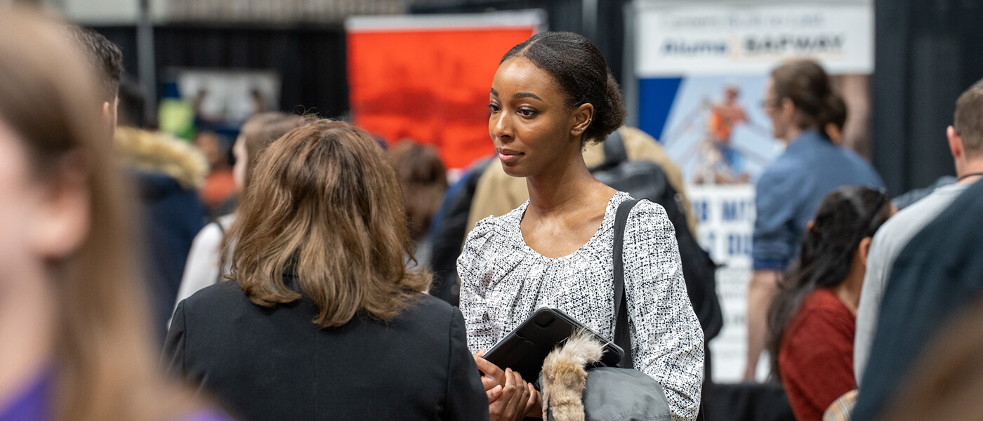 A woman in a patterned blouse holds a coat and notebook, attentively engaging with another person at a busy networking event or job fair.