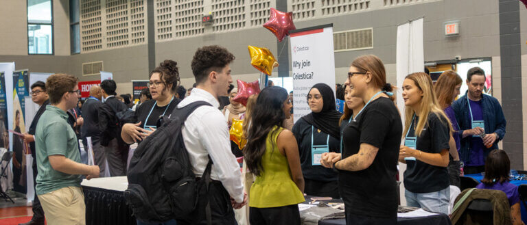 People interact at a bustling job fair, with booths displaying banners. Conversations are lively and engaged, creating an atmosphere of networking and opportunity.