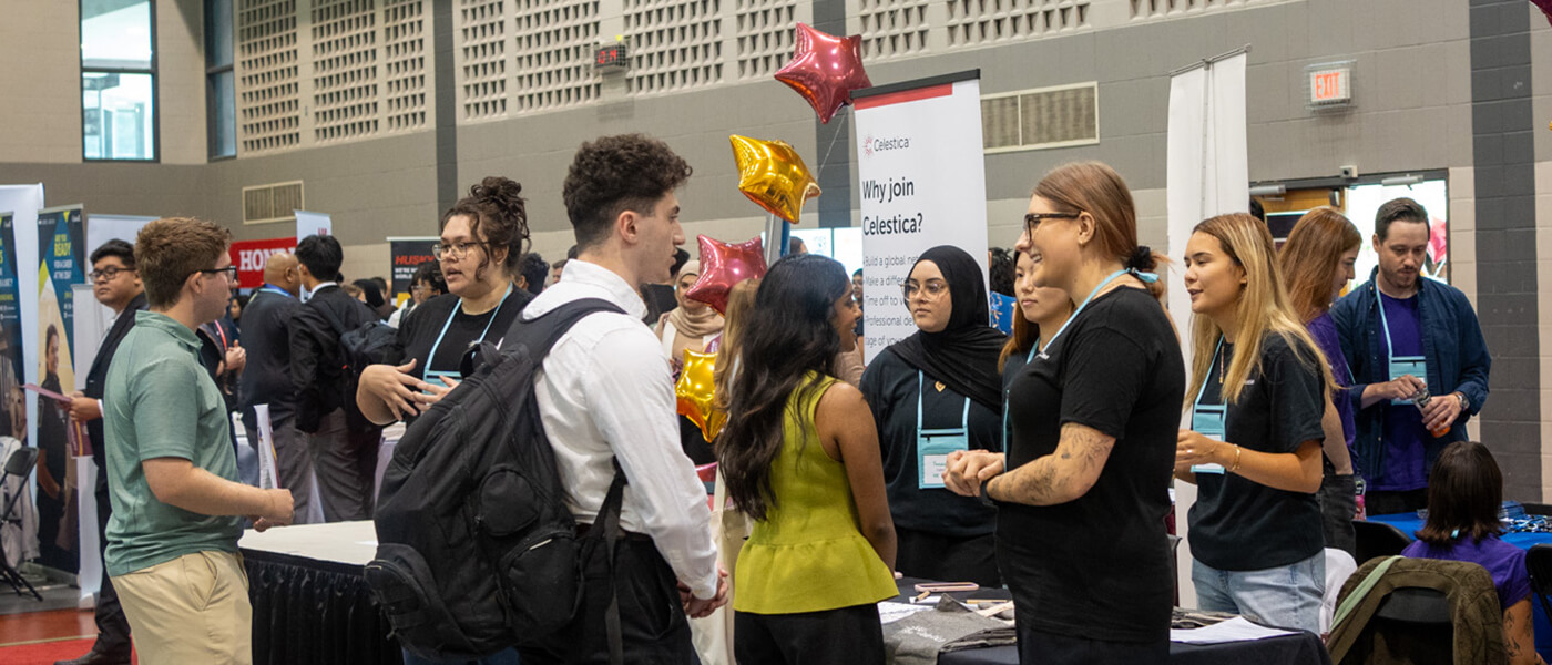 People interact at a bustling job fair, with booths displaying banners. Conversations are lively and engaged, creating an atmosphere of networking and opportunity.