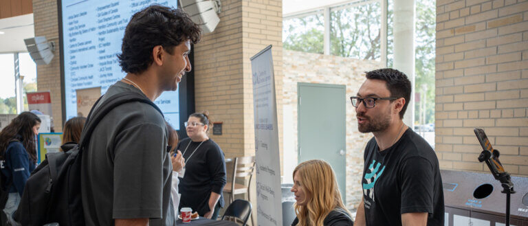 Two men engage in conversation at a college fair. A digital display and event banners are visible. The mood is lively and engaging.