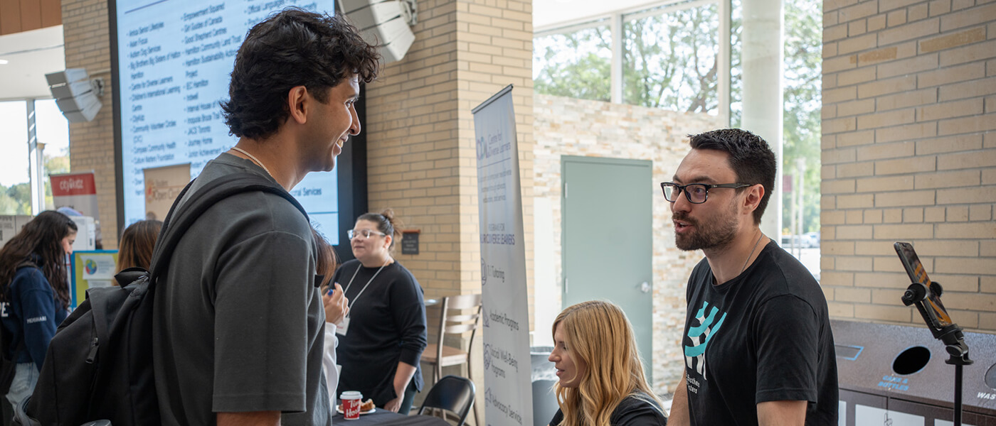 Two men engage in conversation at a college fair. A digital display and event banners are visible. The mood is lively and engaging.
